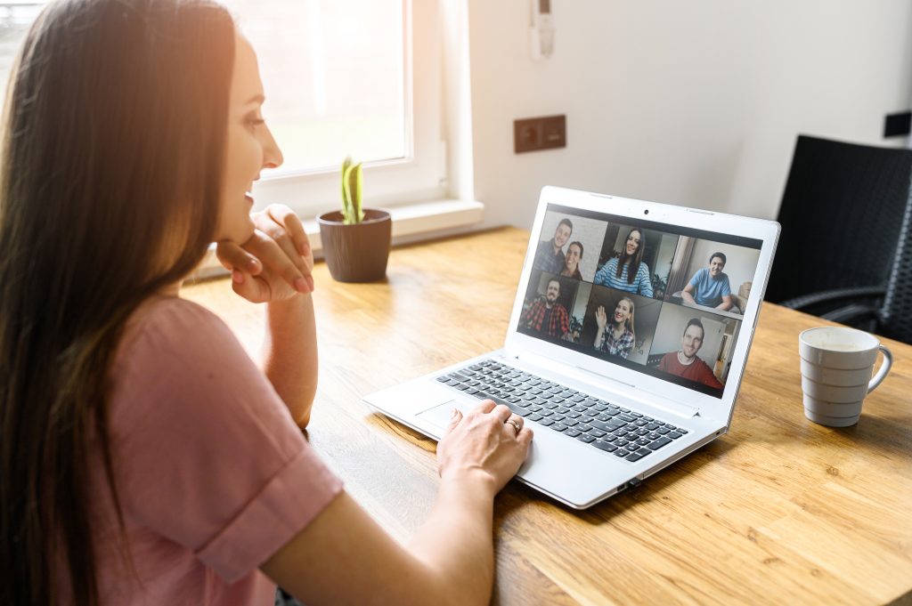 Communication on distance via video, video call, zoom. A young attractive woman using app on laptop for meeting with friends. She looks at webcam and smiles. Side view