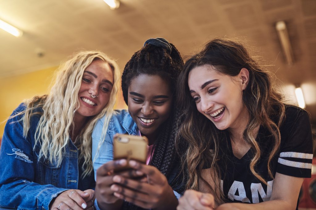 Laughing young female friends leaning on a laundromat counter using a cellphone together while doing laundry